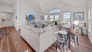 Een ruime woonkamer met een grote L-bank, een houten eettafel met barstoelen, en een keuken met een televisie.