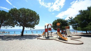 Een speeltuin met glijbanen en klimconstructies, omringd door bomen en een strand met parasols.