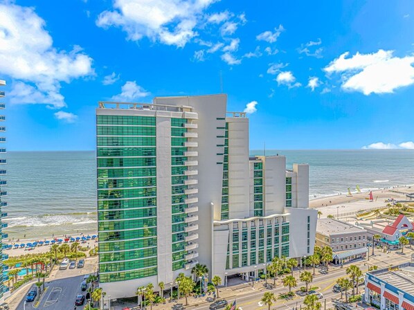 A modern hotel with a prominent glass facade, a beach view, and a parking area.