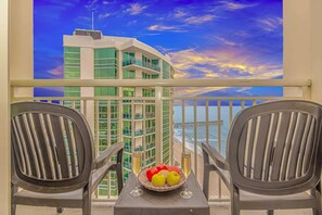 A balcony with two chairs, a table with a fruit bowl and a glass of champagne, and a view of a building and the ocean.