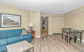 A living room with a blue sofa, a coffee table, a dining table with chairs, and a hallway leading to a bedroom.