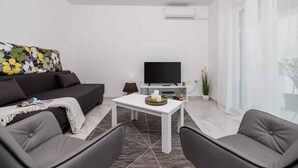 Una sala de estar moderna con un sofá, dos sillones, una mesita de centro, un televisor de pantalla plana y un amplio ventanal con cortinas translúcidas.