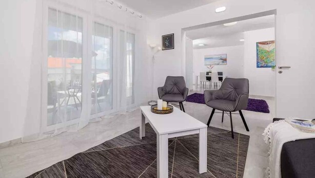 Un salón moderno con una mesa blanca, dos sillones grises y un ventanal con cortinas translúcidas.
