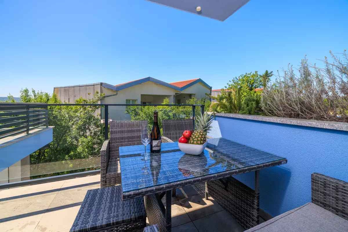 A balcony with a glass table, wicker chairs, a bottle, and a bowl of fruit.