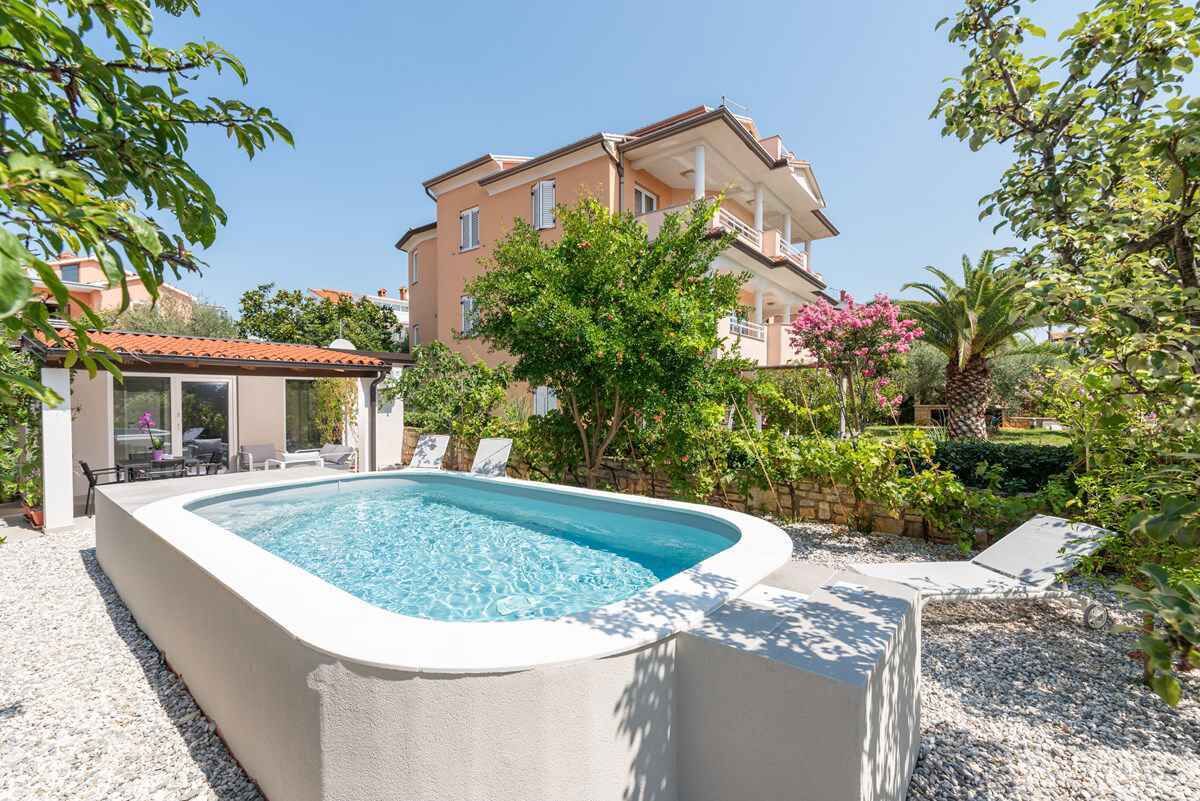 Una piscina con cubierta blanca, rodeada de vegetación y un edificio al fondo.