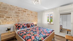 A bedroom with a stone wall, a bed with floral bedding, a nightstand with a lamp, a window with a view of greenery, and a wardrobe.