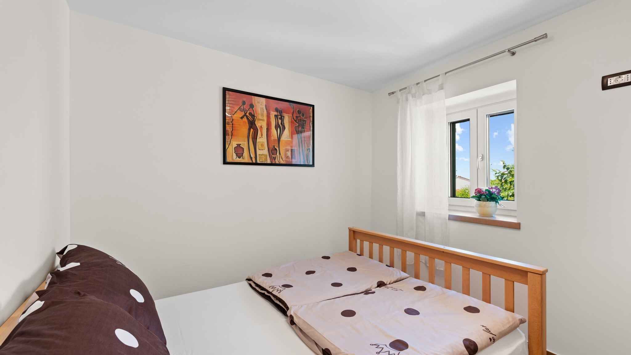 A bedroom with a bed, a window with curtains, a painting on the wall, and a view of greenery outside.