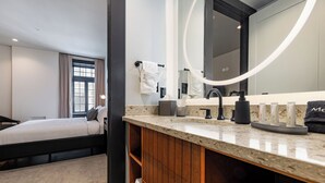 A modern hotel room with a bed, a bathroom with a large mirror, and a vanity with toiletries. - 3BR City Views: Walk to French Quarter | The Mayfair (New Orleans)