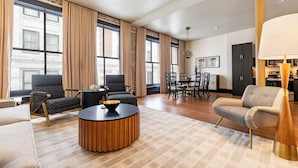 A spacious living room with a modern design, featuring a round wooden coffee table, comfortable seating, and large windows with curtains. - 2BR Elegant French Quarter Escape | The Mayfair (New Orleans)