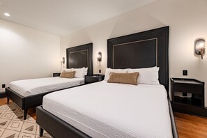 Two single beds with black frames and white bedding, a dark headboard, a small bedside table with a lamp, and a patterned rug on the floor.