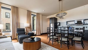 A modern living room with a sofa, a round coffee table, and a dining area with chairs and a table. There is a kitchen area with appliances and a hanging light fixture.