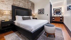 Un dormitorio moderno con pared de ladrillo, una cama grande, una mesita de noche con lámparas, un taburete pequeño y un baño con espejo y estanterías.