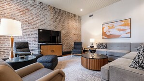 A modern living room with a brick wall, a flat-screen TV, a grey sofa, a round coffee table, and patterned armchairs.