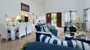 Amplia sala de estar con un sofá azul oscuro, sillas blancas y un comedor con una mesa blanca.