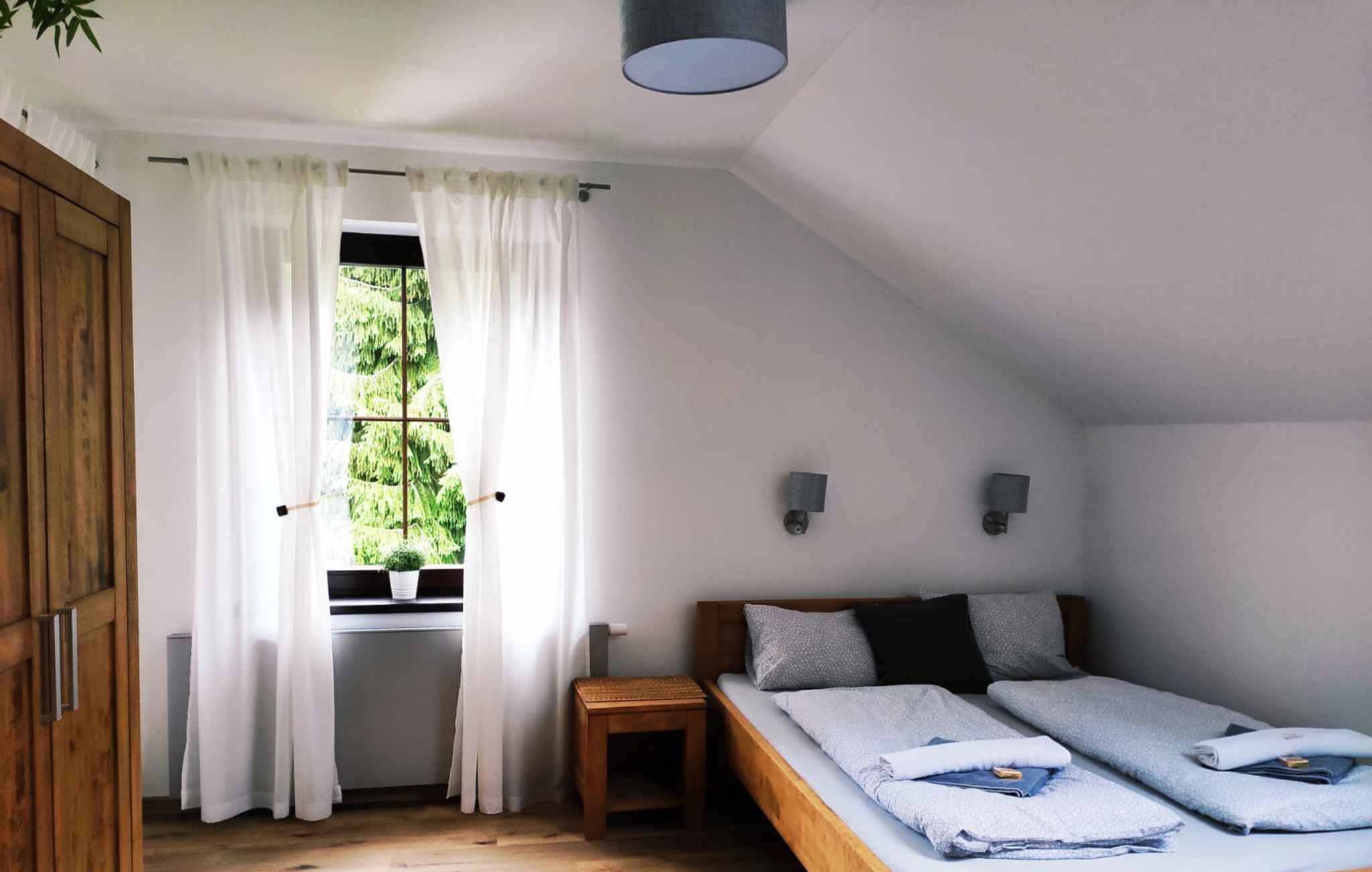 A small, single-room accommodation with a bed, a small table, a window with curtains, and a ceiling light.