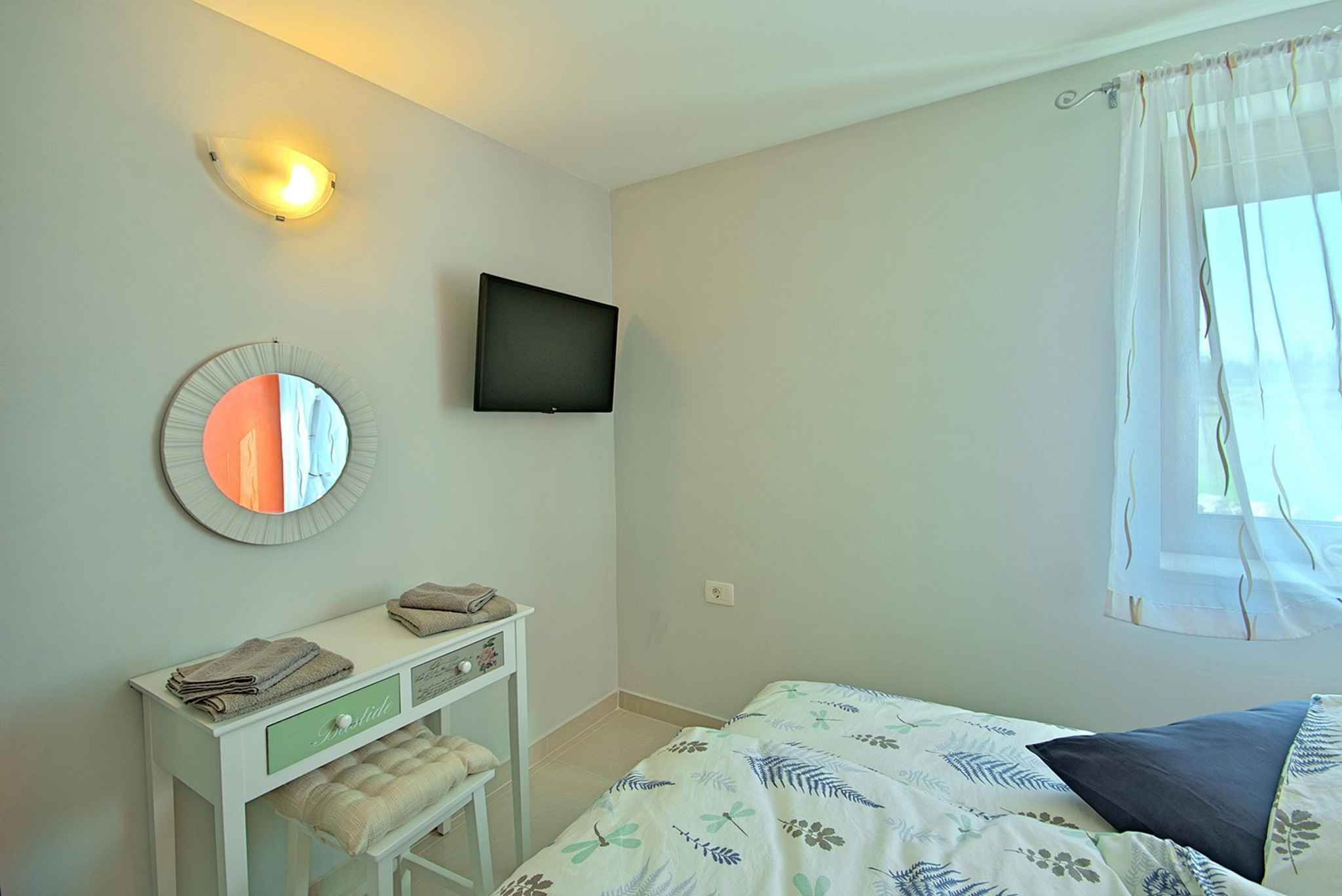 Un dormitorio con cama, televisor montado en la pared, mesita de noche con espejo y ventana con cortinas.