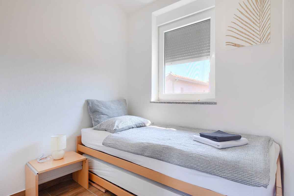 Quarto pequeno com uma cama de solteiro, janela, mesa de cabeceira e prateleira na parede.