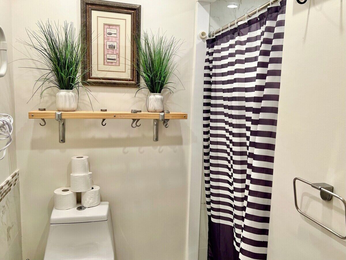 A bathroom with a toilet, two potted plants on a shelf, a striped shower curtain, and a framed picture on the wall.