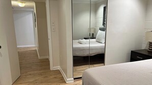 Ein Hotelzimmer mit Holzboden, einem Bett mit weißer Tagesdecke, einer Glastrennwand und einem kleinen Nachttisch mit Lampe.