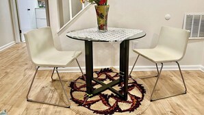 Ein runder Tisch mit einer Blumenvase, zwei Stühlen und einem gemusterten Teppich in einem Raum mit Holzboden und einer Treppe im Hintergrund.