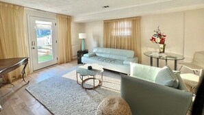 Ein Wohnzimmer mit einem hellblauen Sofa, einem runden Couchtisch, einem Glastisch mit Blumenvase und einer Tür, die auf einen Balkon führt.