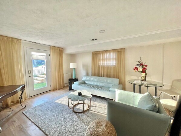 A living room with a light blue sofa, a round coffee table, a glass table with a vase of flowers, and a door leading to a balcony. - 1 Bedroom Basement Apartment | No Car needed (Arlington)