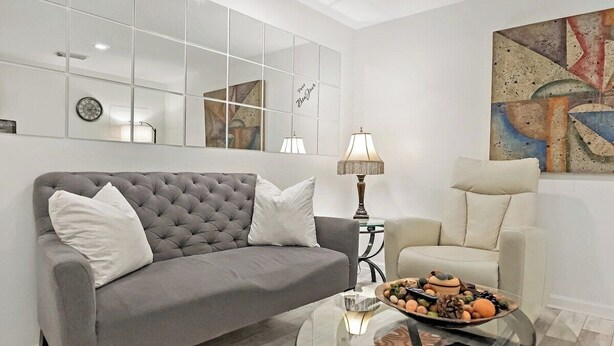 Uma sala de estar com um sofá cinzento, uma poltrona branca, uma mesa de centro de vidro com objetos decorativos, um abajur e um quadro abstrato grande.