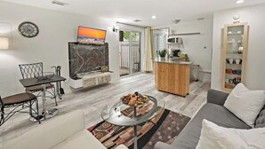 Uma sala de estar moderna com uma mesa de centro de vidro, um sofá, um espaço de jantar com mesa e cadeiras, uma cozinha com micro-ondas e vista para o exterior através de uma porta de vidro.