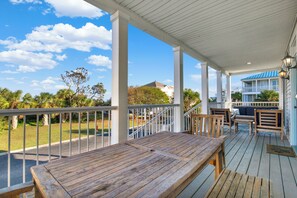 A wooden deck with a table and chairs, overlooking a grassy area and a building with a blue roof. - Seconds to the Sea, Ocean views, Heated Pool Access (Tybee Island)