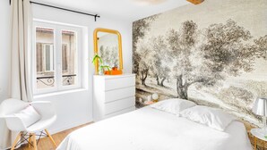 A bedroom with a large bed, a dresser, a chair, a mirror, and a wall with a nature-themed mural. - T2 Bazeilles by Interhome (Toulon)