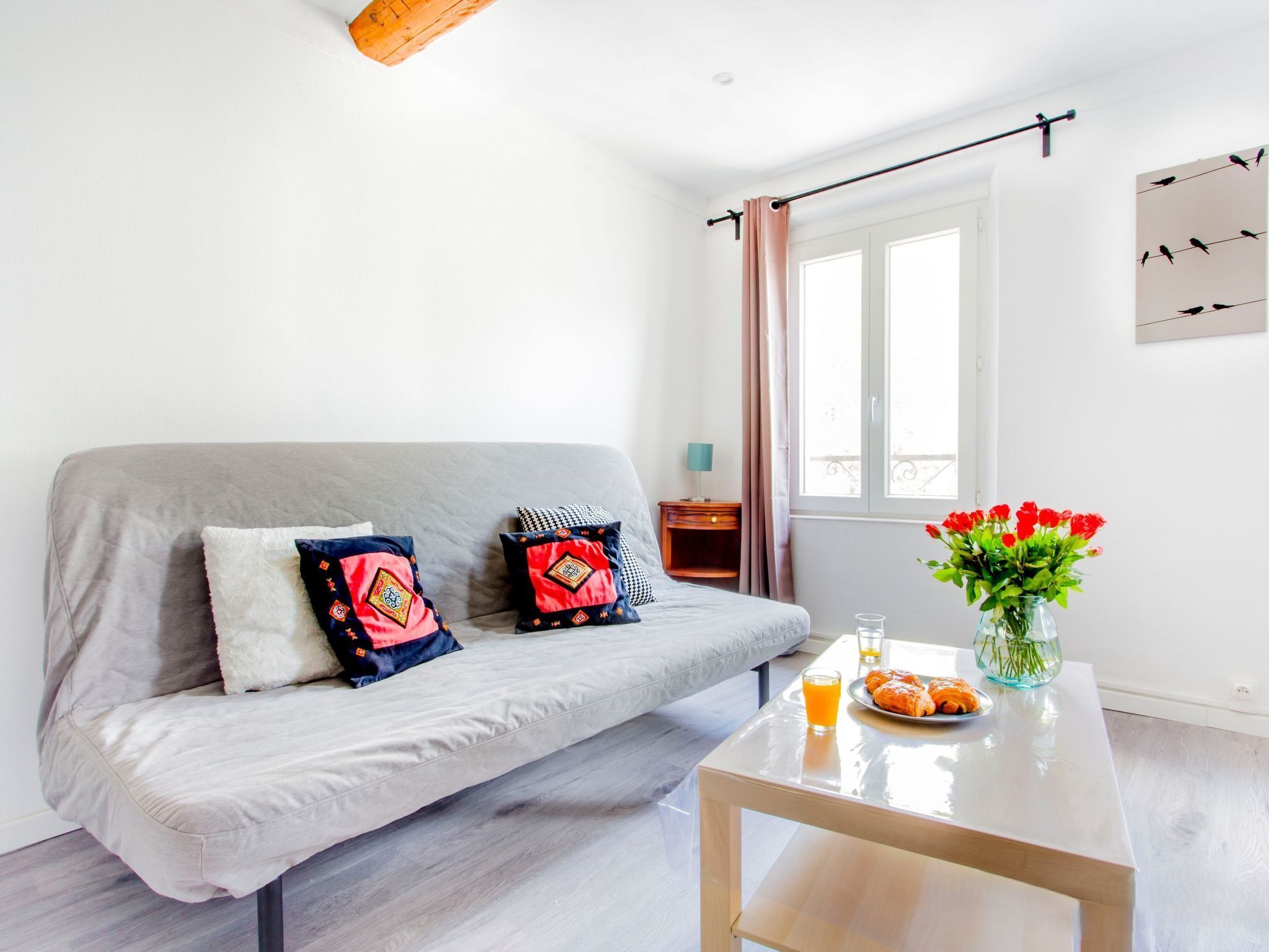 A modern living room with a grey sofa, two decorative pillows, a small wooden side table with a lamp, a glass coffee table with a vase of red flowers, and a breakfast setup with croissants and orange juice.