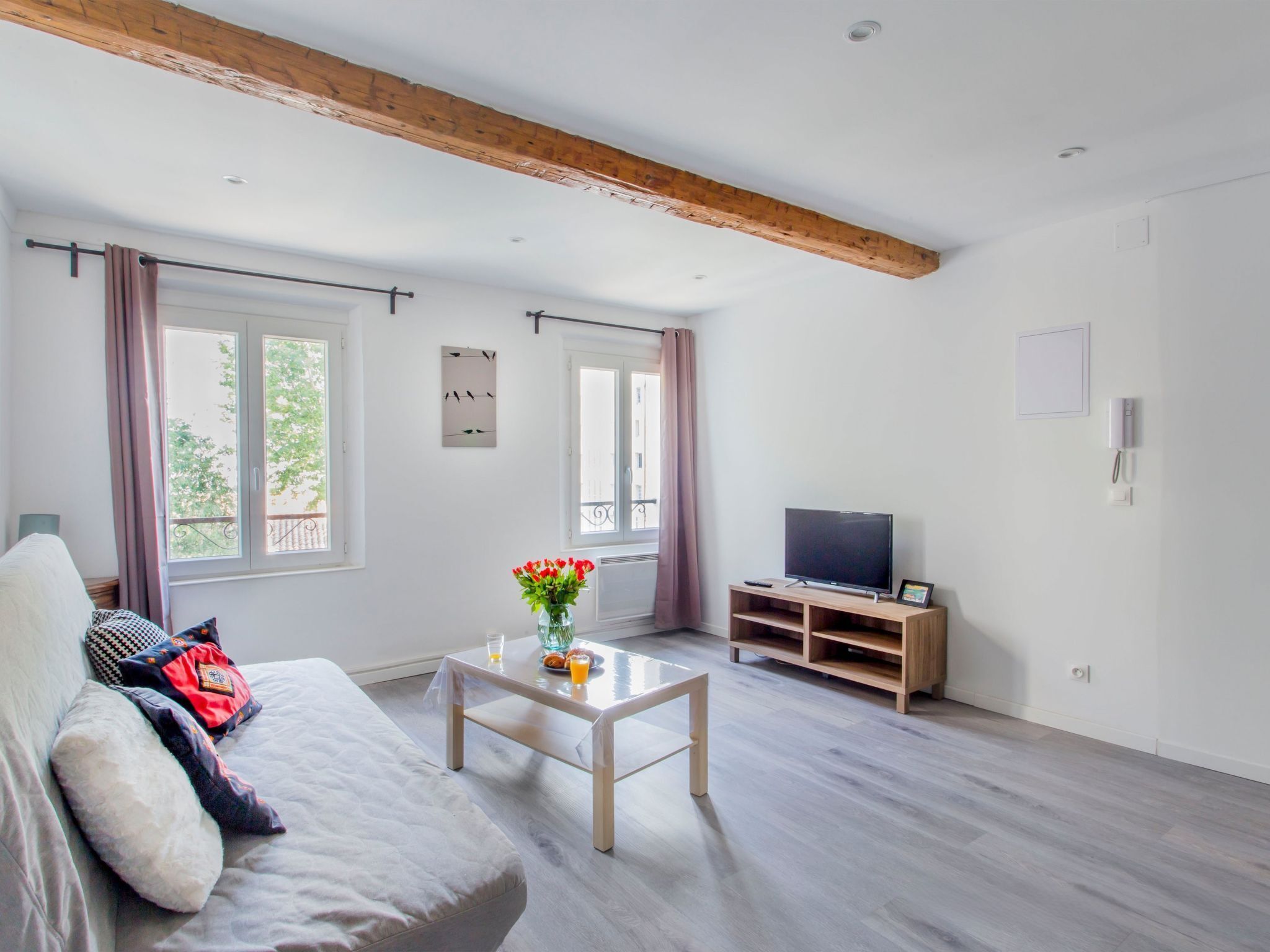 Ein modernes Wohnzimmer mit einer Couch, einem Couchtisch mit Blumen, einem Fernseher auf einem Holzboden und großen Fenstern mit Vorhängen.