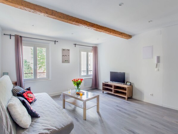 A modern living room with a sofa, a coffee table with flowers, a TV on a wooden stand, and large windows with curtains.
