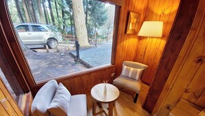 Hyggelig hytteinteriør med sofa, stol og lille bord. Et vindue viser en parkeret bil og et skovområde udenfor.