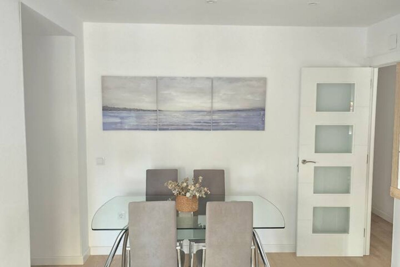 Een eethoek met een glazen tafel, twee stoelen, een vaas met bloemen en een aan de muur hangend schilderij.