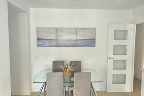 A dining area with a glass table, two chairs, a vase with flowers, and a wall-mounted painting.