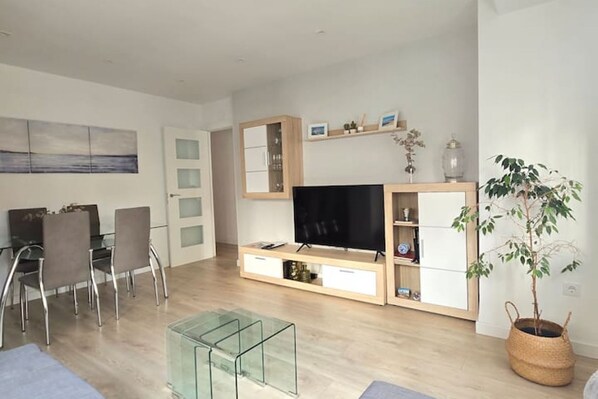 A modern living room with a flat-screen TV, glass coffee table, dining area with chairs, and a potted plant.