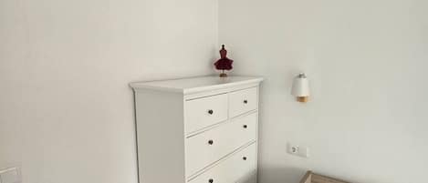 A white dresser with drawers, a small red object on top, and a wall-mounted light fixture.