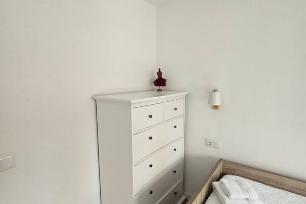 Un aparador blanco con cajones, un objeto rojo pequeño encima y una lámpara de pared.