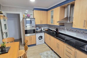 Cocina compacta con armarios de madera, horno integrado, lavadora y comedor con sillas y mesa.