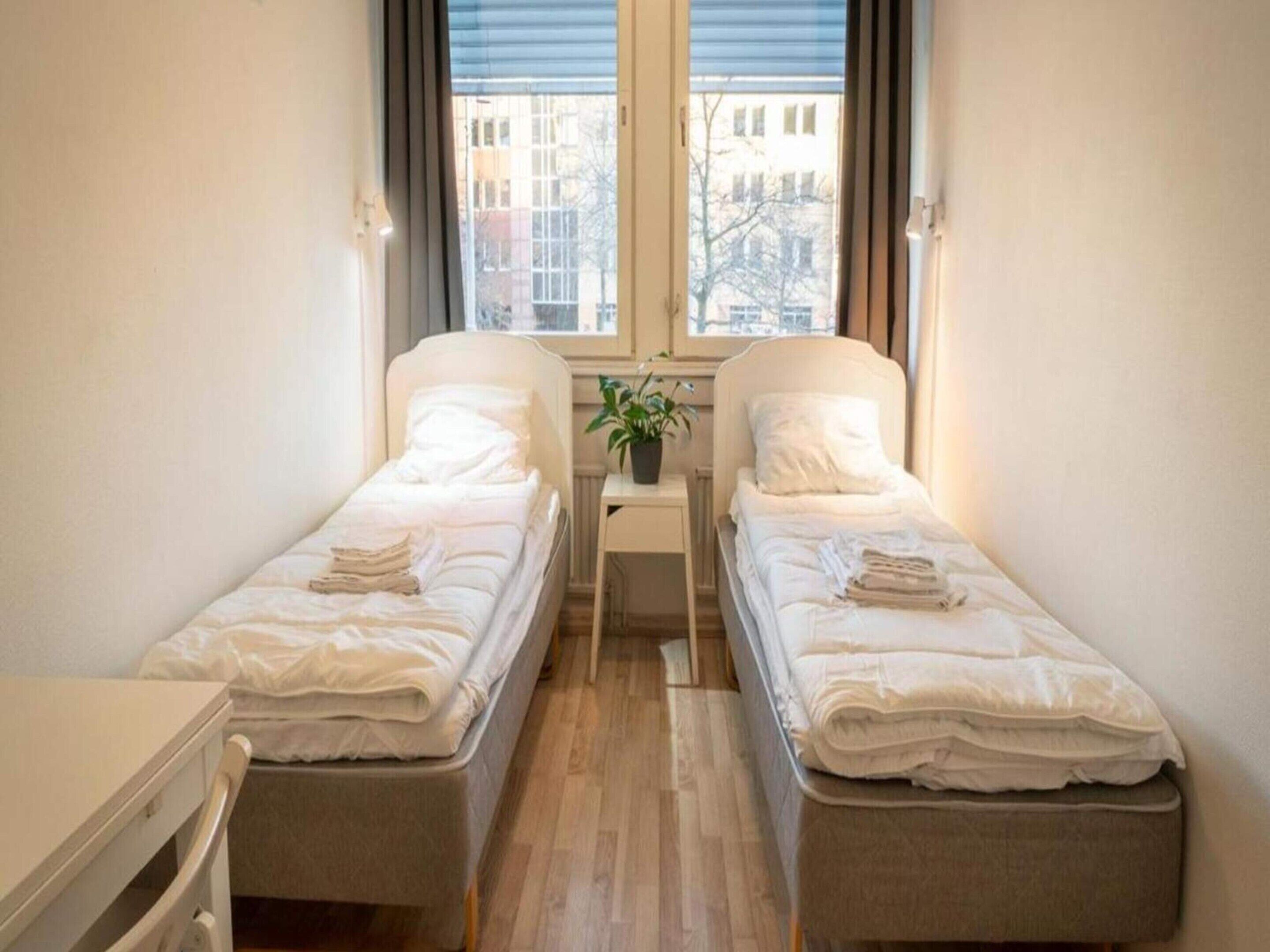 Ett litet, rent rum med två enkelsängar, ett litet bord med en växt och ett fönster med utsikt över byggnaderna utanför.
