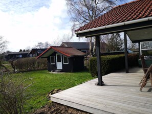 Eine Holzterrasse führt zu einem kleinen Haus mit rotem Ziegeldach, umgeben von viel Grün und weiteren Häusern.