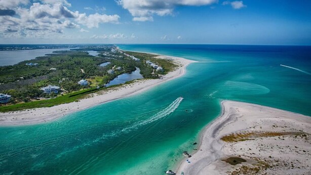 Ein Luftbild einer Küstenlandschaft mit Strand, klarem türkisblauem Wasser und einer kleinen Siedlung mit Häusern und Grünflächen.