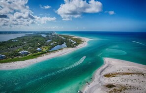 An aerial view of a coastal area with a beach, clear turquoise waters, and a small settlement with houses and greenery.