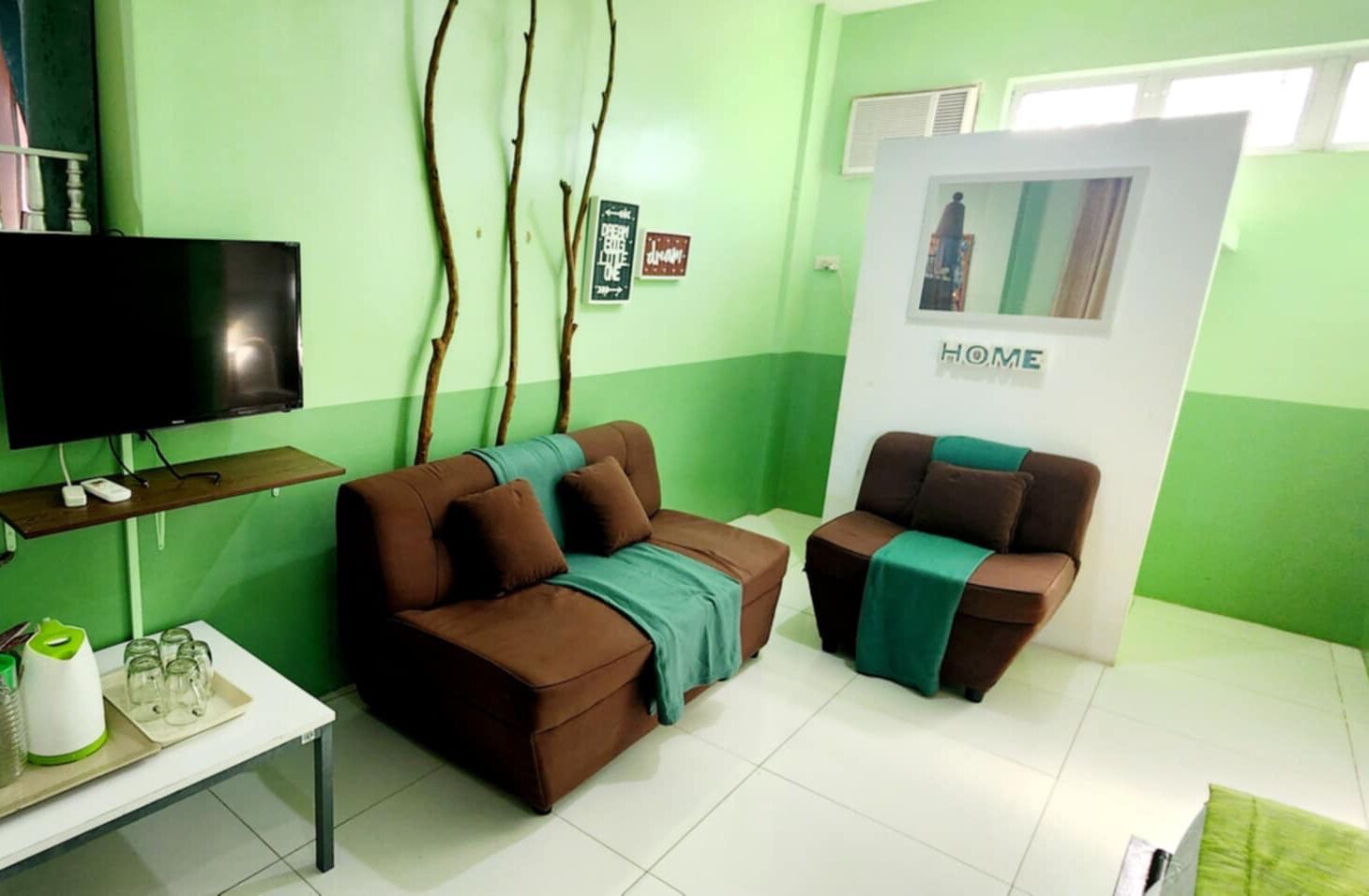 A living room with two brown sofas, a flat-screen TV, a small table with glasses, and a 'HOME' sign on the wall.