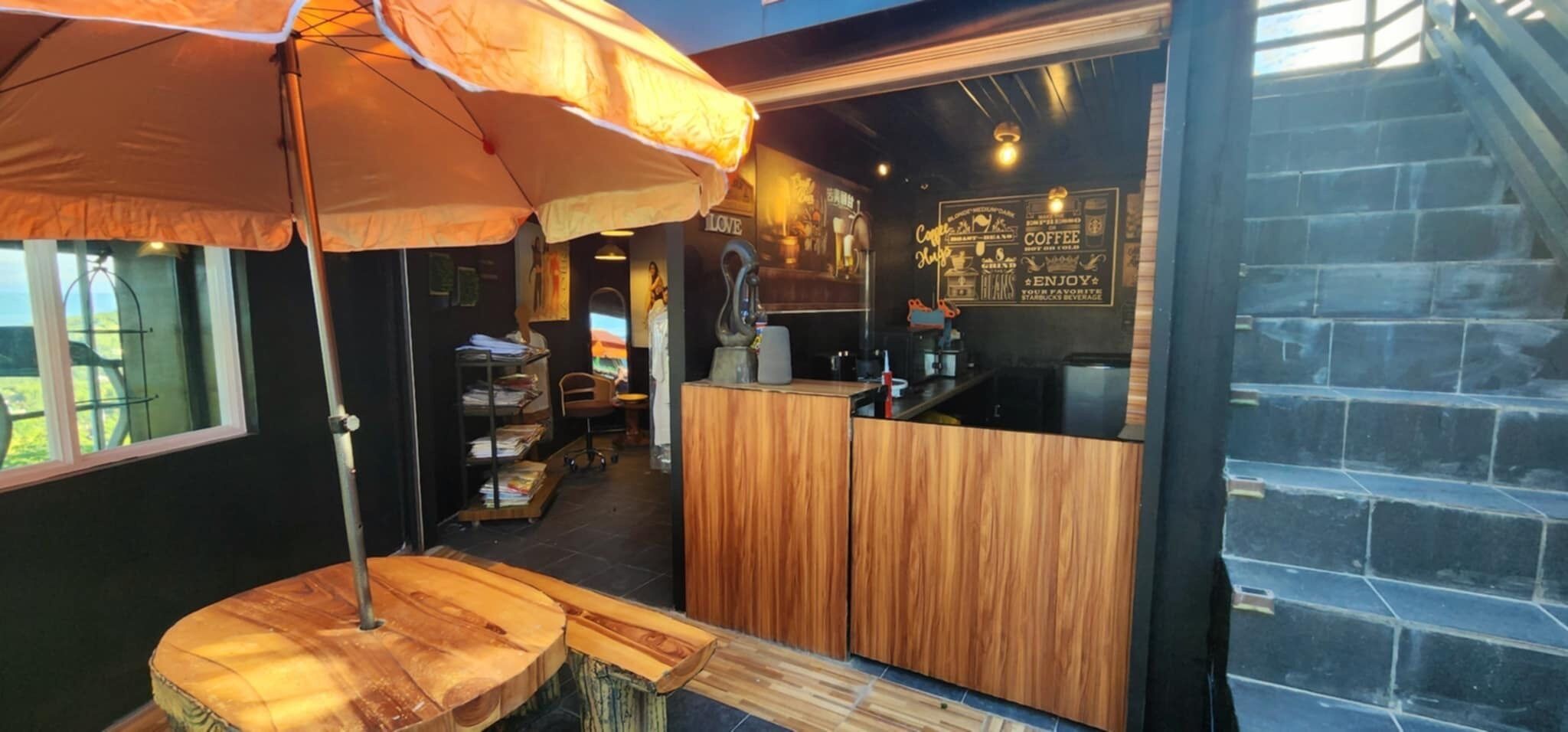 A cozy coffee shop interior with a wooden counter, a large umbrella, and a staircase leading upstairs.