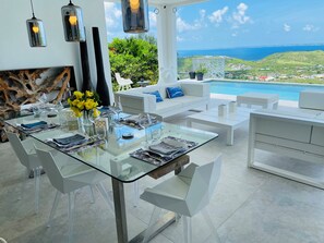 A modern outdoor dining area with a glass table, white chairs, and a view of the ocean.
