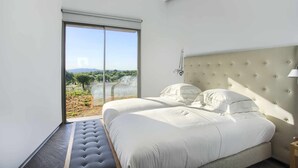 Ein Schlafzimmer mit einem großen Bett, einem gepolsterten Kopfteil und Blick auf eine Landschaft durch eine Schiebetür.
