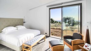 Une chambre moderne avec un grand lit, un banc, une chaise et une vue sur l’extérieur grâce à une porte-fenêtre coulissante.
