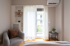 Una stanza con una poltrona grigia, un tavolino con una pianta, uno scaffale bianco con oggetti decorativi e una porta scorrevole in vetro che dà su un balcone esterno.